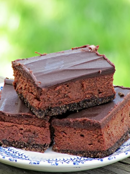 A sdie view of three rich chocolate chocolate cheesecake rectangles with chocolate cookie crust, chocolate cream cheese filling and a dark chocolate glaze on top, stacked on a blue and white plate aginst a green background.