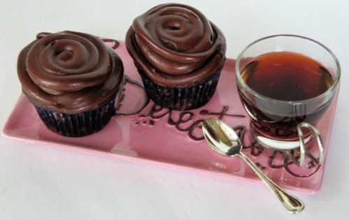 Overhead view of a rectangular pink tray with brown writing on it holding two large chocolate cupcakes with chocolate espresso frosting mounded high on the cupcakes, along with a cup of black coffee in a glass cup and a spoon.