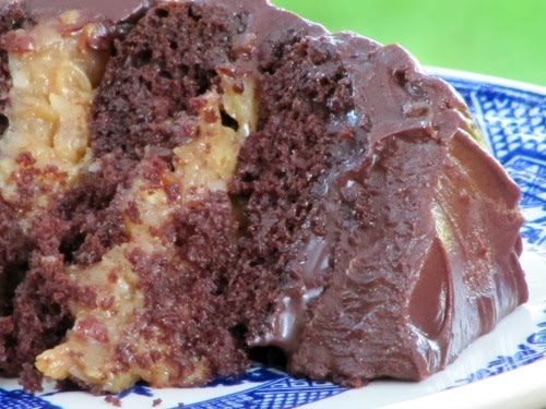 closeup of a slice of rich coconut-pecan filled inside out german chocolate cake with chocolate frosting is sitting on its side on a blue and white plate against a green background.