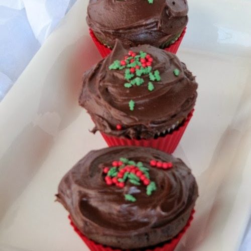 CLoseup view of three chocolate gingerbread cupcakes in red liners on a white tray with dark chocolate Hershey's frosting on their tops and red and green sprinkles.