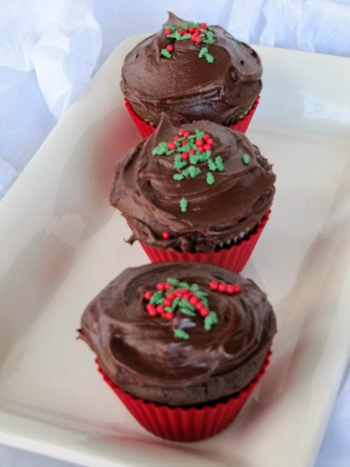 CLoseup view of three chocolate gingerbread cupcakes in red liners on a white tray with dark chocolate Hershey's frosting on their tops and red and green sprinkles.