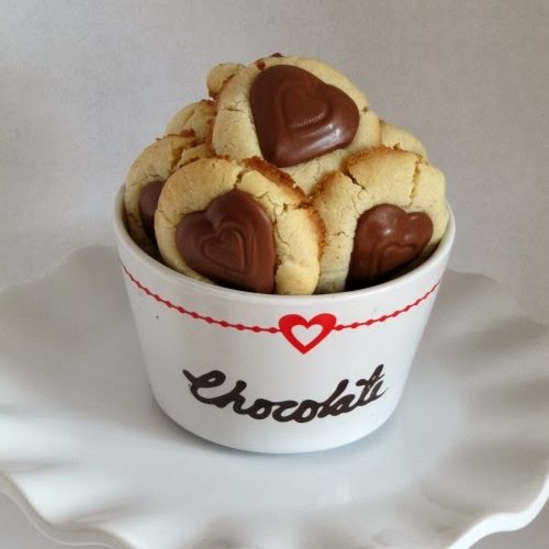 A fluted white platter holds a white dish of round shortbread valentine peanut butter heart thumbprint cookies with chocolate penut butter-filled heart shaped candy centers. The dish says chocolate in black script and has a red heart decorating the upper edge.