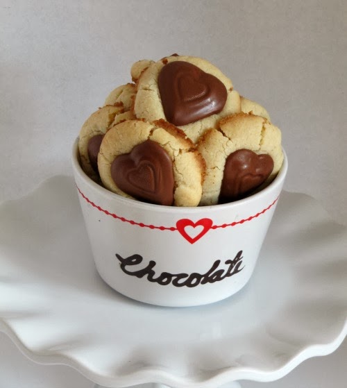 A fluted white platter holds a white dish of round shortbread valentine peanut butter heart thumbprint cookies with chocolate peanut butter-filled heart shaped candy centers. The dish says chocolate in black script and has a red heart decorating the upper edge.
