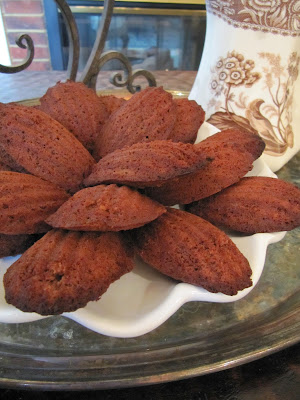 A full plate of Chocolate Madeleines or Madeleines au Chocolat sitting on a round table in front of a white pitcher with tan floral designs.