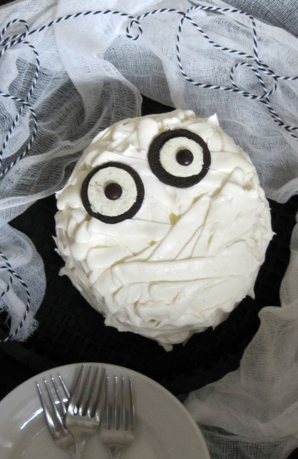Overhead view of a three-layer chocolate cake with vanilla buttercream frosting decorated to look like a chocolate mummy cake for your halloween celebrations with two Oreo halves and 2 brown M&Ms for eyes, set against some white cheesecloth and blue and white string and white dessert plates.