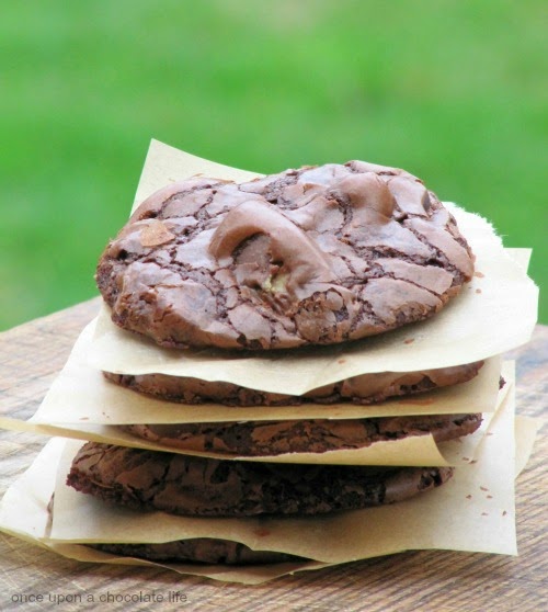 A closeup image of rich chocolate reese's peanut butter egg cookies with crackled tops in a stack, separated with squares of light brown parchment paper on a wood surface against a green background.