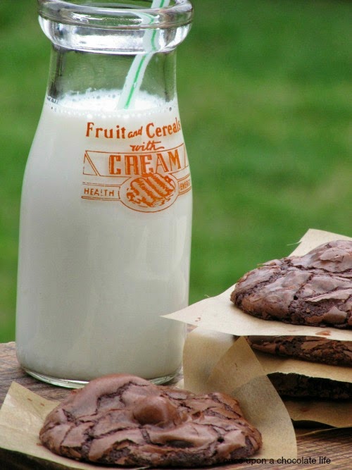 A stack of chocolate Reese's peanut butter egg cookies and a single chocolate cookie are sitting on a brown surface in front of a glass milk bottle filled with milk against a green background.