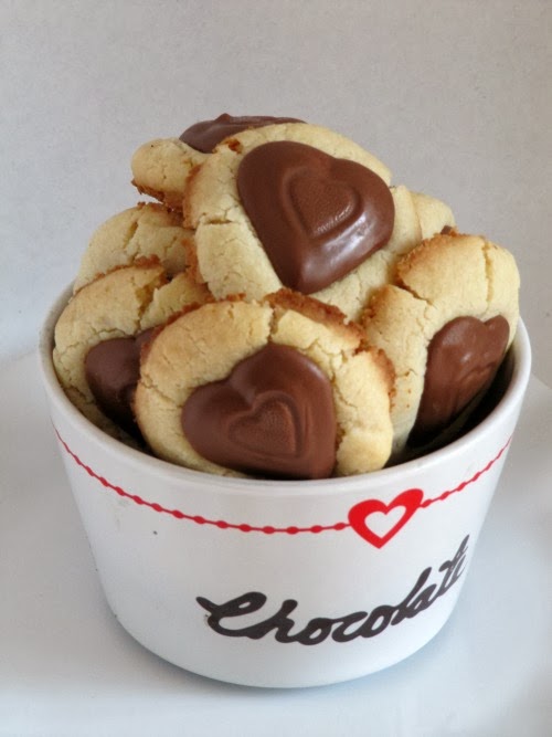 A white bowl with the word 'chocolate' on it and a red heart holds several valentine peanut butter heart thumbprint cookies made with shortbread and Reese's peanut butter heart candies.