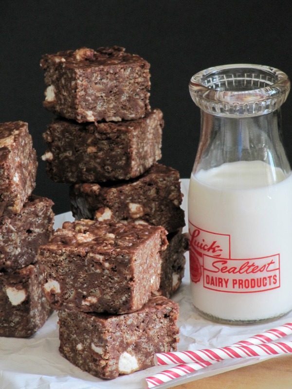 A large stack of chocolate peanut butter marshmallow treats are sitting on a white table cloth next to a glass milk bottle half full of milk and two red and white straws on a wood table and against a black background.
