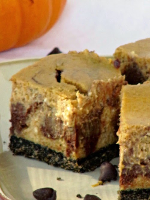 A side closeup view of three chocolate pumpkin cheesecake bars with a graham cracker and oreo crust and a pumpkin cheesecake filling with chocolate swirls are sitting on a white plate with an orange pumpkin in the background.