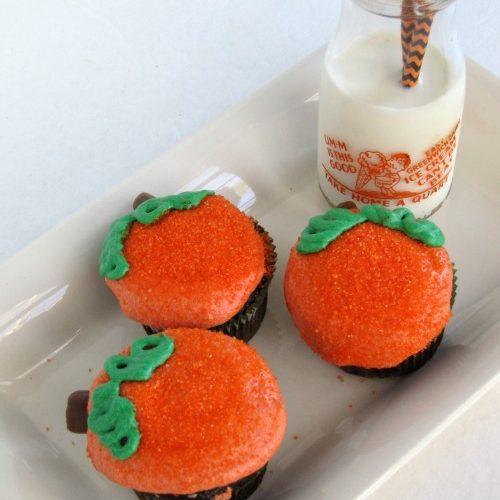 Overhead view of a white rectangular tray against a white background holding three chocolate cupcakes with orange buttercream frosting and green leaf frosting decoration along with a glass milk bottle half full of milk and two halloween colored straws.