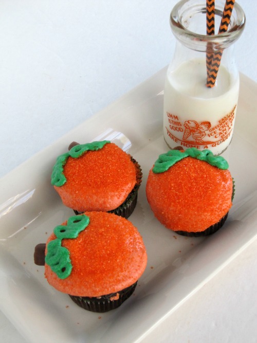 Overhead view of a white rectangular tray against a white background holding three chocolate cupcakes with orange buttercream frosting and green leaf frosting decoration along with a glass milk bottle half full of milk and two halloween colored straws.