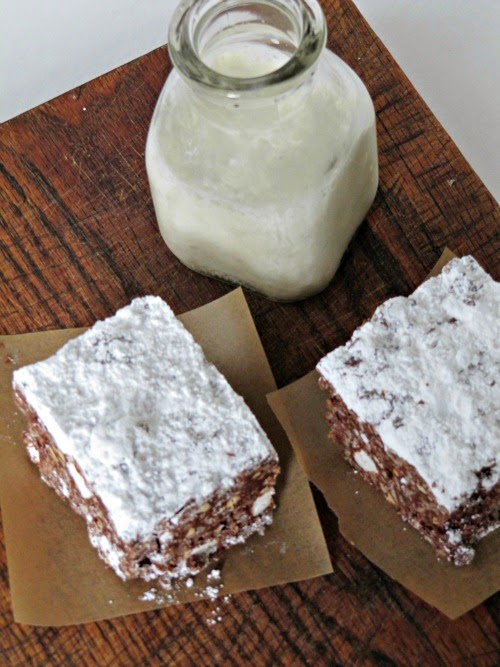 Overhead view of two puppy chow krispie treats that include peanut butter, chocolate, marshmallows and rice krispies and are dusted with powdered sugar sitting on pieces of brown parchment on a wood surface along with a glass bottle of milk.