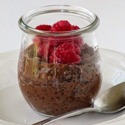 A glass of creamy chocolate tapioca pudding with a fresh red raspberry garnish is sitting on a white plate along with a silver spoon against a white background.