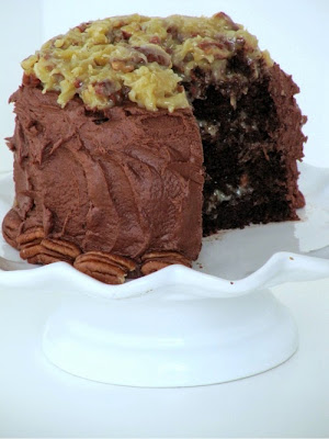 A white footed cake stand holds a large round Chocolate Velvet Cake with Fudge Filling and Coconut Pecan Frosting that shows a piece cut out of it revealing the innder layers of the cake.