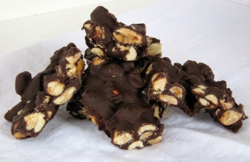 A pile of salted caramel peanut bark chunks containing large peanut pieces, salted caramel and covered in dark chocolate are sitting on a white surface against a white background.