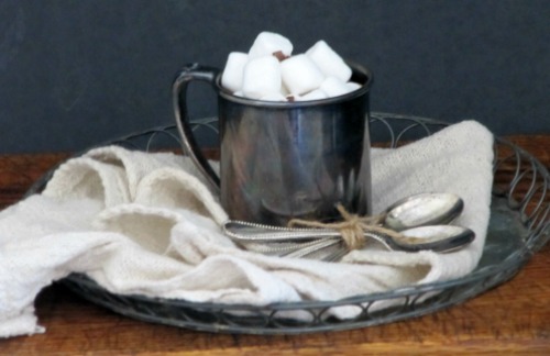 Side view of a dark gray mug filled witih real simple magazine hot cocoa mix hot chocolate and topped with melted marshmallows. The mug is against a dark background on a white cloth on a silver tray along swith three silver spoons.