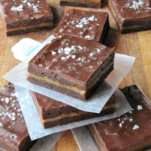 Several square-cut Connie's caramel-stuffed sea salt brownies are scattered across a brown wood table.
