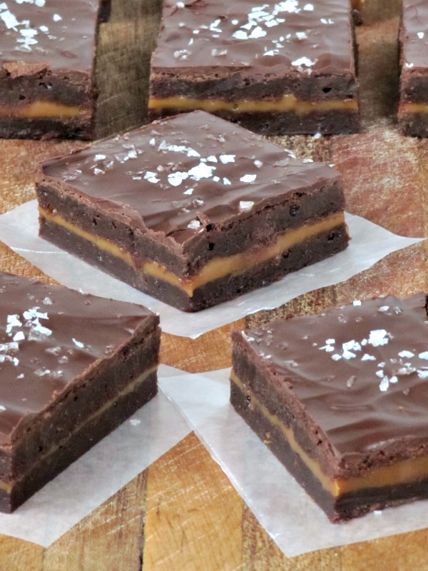 A closeup view of several layered Connie's Caramel-stuffed sea salt brownies with a layer of caramel in between two brownie layers topped with milk chocolate glaze and a sprinkle of sea salt sitting on a wood table.