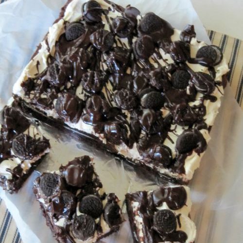 An overhead view of a square block of cookies and cream brownies along with three separate cut brownies made with a brownie base, layer of Oreos, fluffy marshmallow frosting, fudge glaze and mini oreos. The brownies are sitting on a piece of parchment against a blue and white striped cloth.