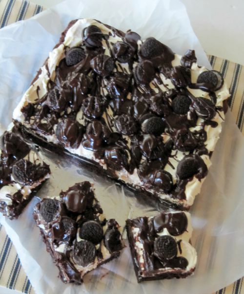An overhead view of a square block of cookies and cream brownies sitting on a piece of parchment against a blue and white striped cloth along with three separate cut cookies and cream brownies.
