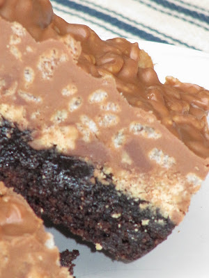 A closeup angled view of a peanut butter cup crunch brownie with a chewy fudgy brownie base topped with melted reese's peanut butter cups, peanut butter, rice krispies and peanuts.