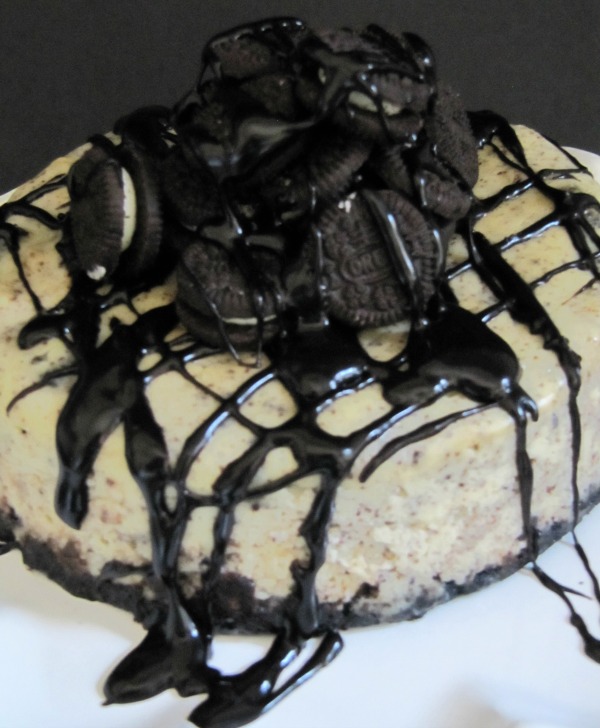 A close up image of a large round oreo fudge baked cheesecake with an oreo crust, crushed oreo and cheesecake center and garnished with a large pile of whole oreo cookies in the middle of the top of the cheesecake with chocolate fudge sauce drizzled all over the top of the cake.