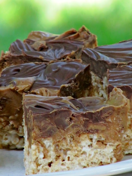 A closeup side view of thick double peanut butter chocolate rice krispie treat squares with white rice krispie bases and thick chocolate chip and peanut butter reeses's candy swirls on top are on a white plate against a green background.