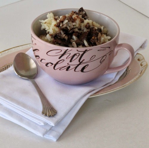 A large pink mug is shown sitting on a white napkin on a pink plate and is holding a serving of chocolate almond toffee rice pudding made with cooked rice, cream, sugar and a chocolate toffee candy bar.
