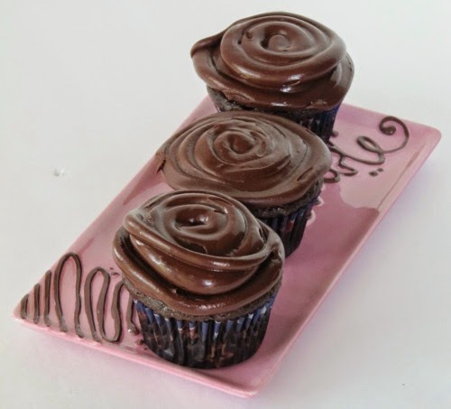 Angled view of three large chocolate cupcakes with chocolate espresso frosting on a pink tray against a white background.