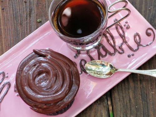 Closeup overhead view of a chocolate cupcake with chocolate espresso frosting swirled on the top in a rich, glossy mound, sitting on a pink ceramic tray along with a cup of black coffee and a silver spoon on a wood table.