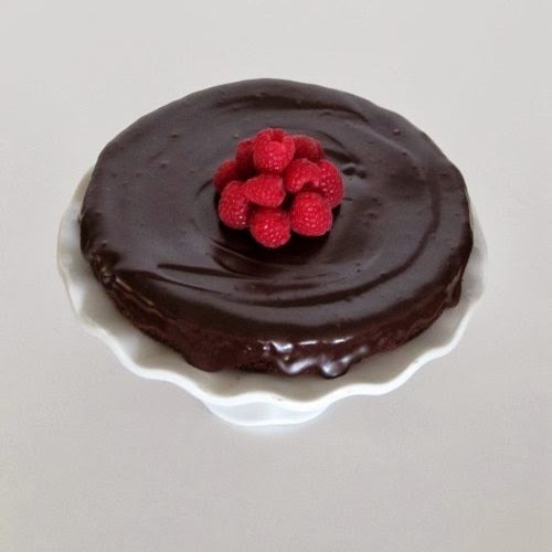 A top view of a white cake stand holding a round glistening flourless chocolate cake glazed with shiny chocolate ganache and with a mound of red fresh raspberries in the center of the cake.