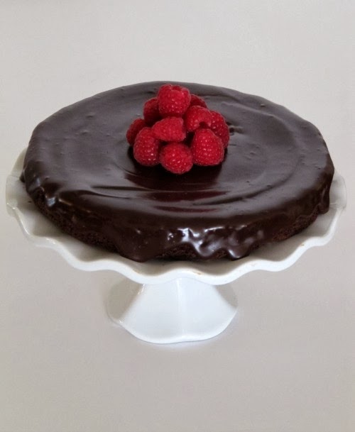 A white footed cake stand shown against a light gray background holds a round flourless chocolate cake with ganache topping with a mound of fresh raspberries in the center of the cake.