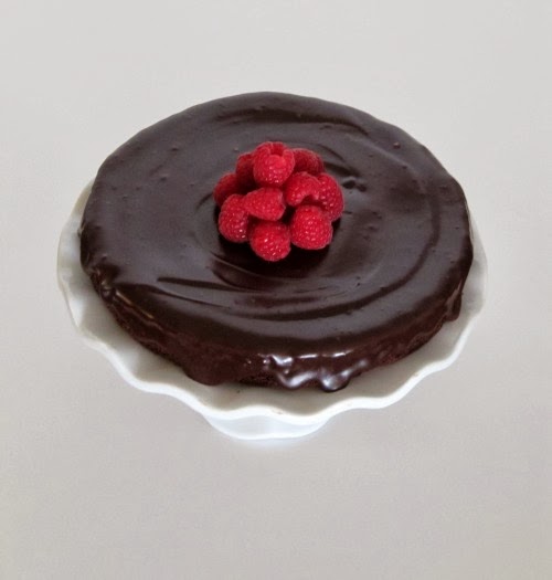 A top view of a white cake stand holding a round glistening flourless chocolate cake with chocolate ganache frosting and with a mound of red fresh raspberries in the center of the cake.