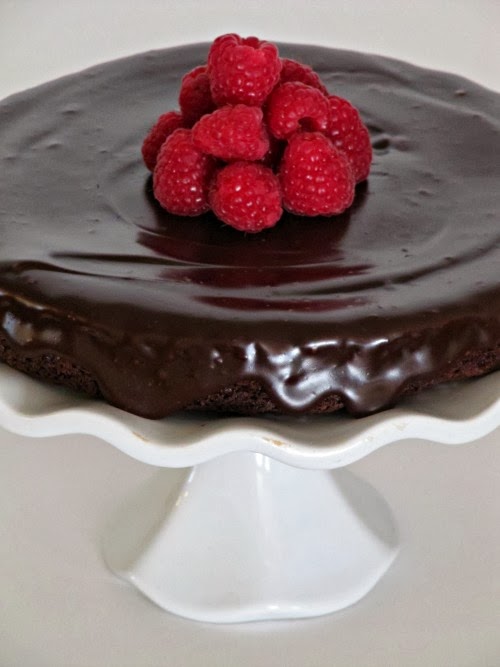 A closeup view of a white footed cake stand holding a round 4-ingredient flourless chocolate cake with chocolate ganache frosting and a center garnish of fresh red raspberries.