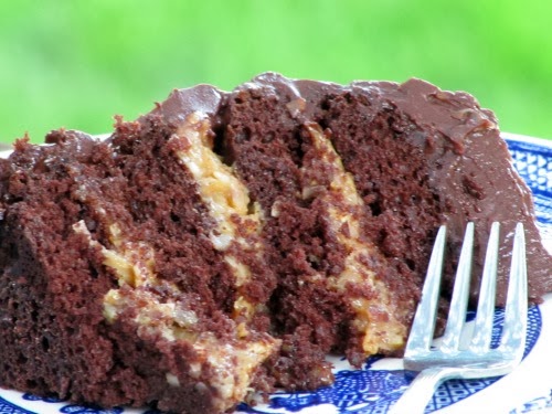a thick slice of inside out german chocolate cake with caramel coconut pecan filling covered with fudge frosting sits on a blue and white plate with a silver fork against a green background.