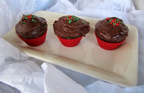 A horizontal view of three Chocolate Gingerbread Cupcakes with Hershey's Perfectly Chocolate Frosting sitting on a white tray on top of white crushed fabric. The cupcakes are in red cupcake liners and are topped with red and green sprinkles on the frosting.