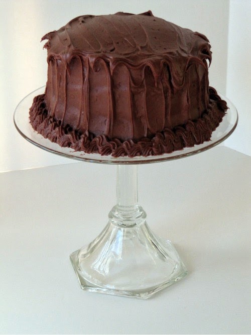 A tall glass cake stand is holding a round Hershey's perfectly chocolate cake frosted with Hershey's perfectly chocolate frosting.