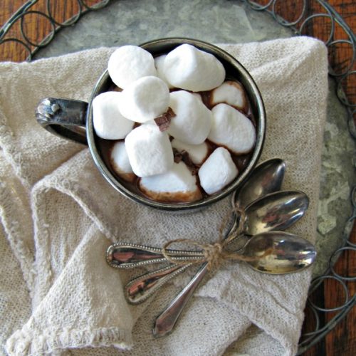 overhead view of an antique mug filled with hot chocolate made with real simple hot cocoa mix and topped with a pile of mini-marshmallows sitting on a white napkin with three silver spoons on a round metal tray