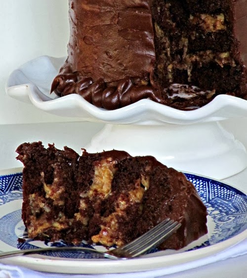 A slice of inside out german chocolate cake sits on a blue and white plate with a fork in front of a white cake stand holding a round chocolate frosted german chocolate cake