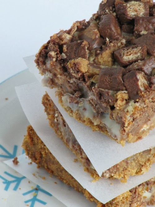 An angled view of a stack of chocolate peanut butter chunk magic bars separated by white parchment squares and showing the rich graham cracker and sweetened condensed milk layers, along with the peanut butter cup and kit-kat candy layer on top.