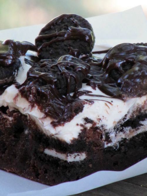 A closeup of a cookies and cream brownie made of a dark chocolate brownie base, a layer of Oreo cookies, marshmallow frosting and a chocolate fudge drizzle on top with mini Oreos.