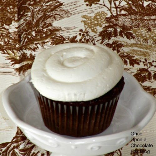 A single chocolate cupcake frosted with marshmallow buttercream frosting for chocolate cupcakes is sitting on a white cake stand against a brown, tan and beige vintage floral cloth.