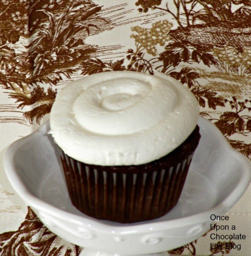 A single chocolate cupcake frosted with marshmallow buttercream frosting for chocolate cupcakes is sitting on a white cake stand against a brown, tan and beige vintage floral cloth.