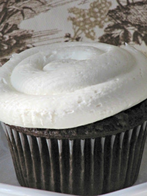 A closeup photo of marshmallow buttercream frosting for chocolate cupcakes is topping a dark chocolate cupcake shown against a brown and white vintage floral background.