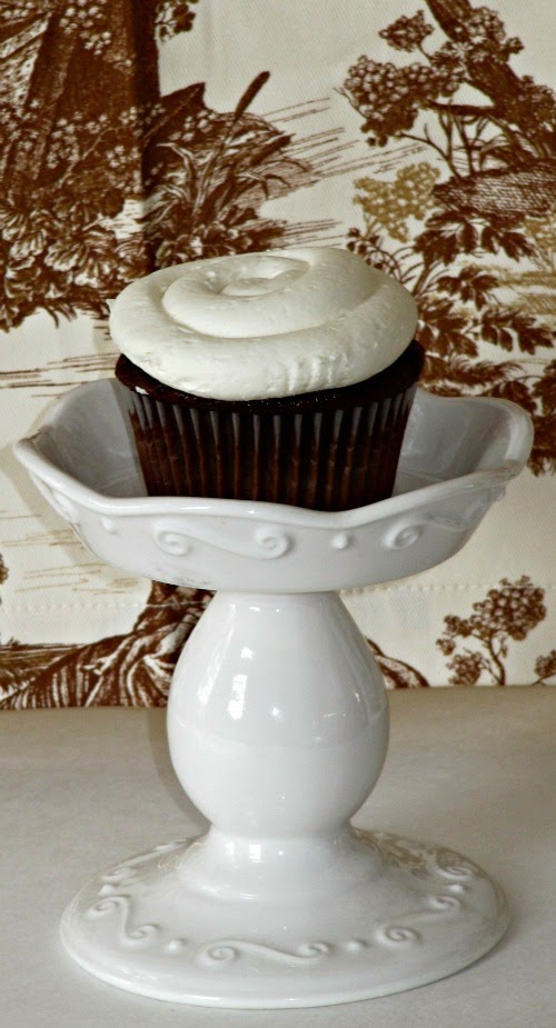 A single large dark chocolate cupcake with marshmallow buttercream frosting is sitting in a footed white ceramie stand against a brown and tan floral background.