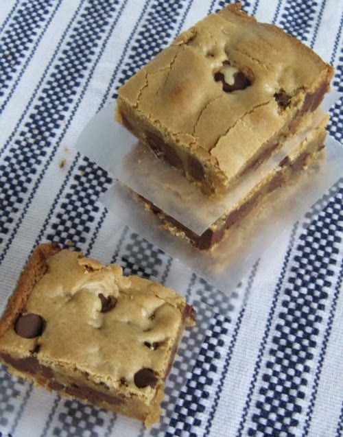 Overhead view of two stacked triple chocolate blondie bars with one to the side, filled with milk, semi-sweet, and dark chocolate chips on a blue and white striped woven table cloth.