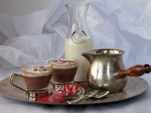 a front view of a crumpled white background against which a silver tray holds two mugs of Neilsen Massey Peppermint Hot Chocolate with whipped cream topping and crushed peppermints with a glass bowl of peppermint candy, silver spoons, a silver pot of hot chocolate and a glass pitcher of milk.