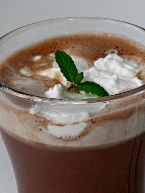 very close view of the top of a glass mug of  basil mint hot chocolate drink with a whipped cream garnish and a sprig of fresh mint.