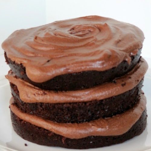 A side view of a three-layer moist chocolate cake and fudge buttercream frosting in between the layers and on top, sitting on a white plate against a white background.